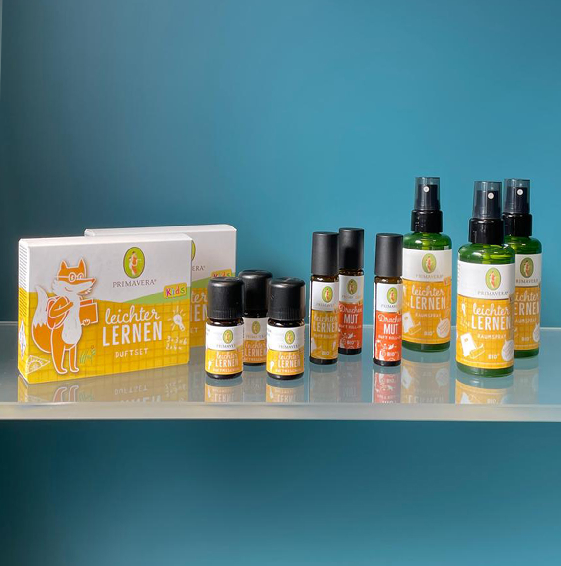 Mehrere Primavera Aroma-Roll-Ons und Raumsprays für Kinder auf einem transparenten Regal vor einer blauen Wand.