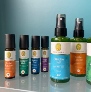 Eine Nahaufnahme zeigt vier Aroma-Roll-Ons und zwei grüne Raumspray-Flaschen der Marke Primavera mit bunten Etiketten, die nebeneinander auf einer spiegelnden Glasfläche stehen.