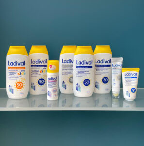 Sonnenschutz & Hautpflege (A) Das Produktfoto zeigt eine Auswahl an Ladival-Sonnenschutzprodukten für verschiedene Hauttypen – von Kinderhaut über allergische bis hin zu trockener Haut – aufgereiht auf einer spiegelnden Glasfläche vor blauem Hintergrund.