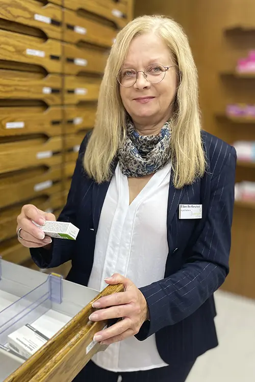Ellen Reifferscheid steht an einer geöffneten Schublade, hält ein Medikament in der Hand und lächelt in die Kamera.