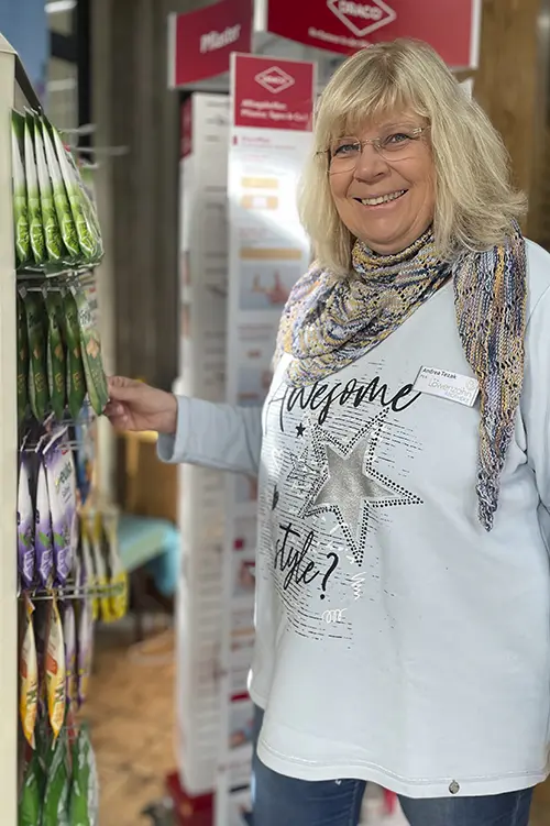 Andrea Tezak steht neben einem Regal mit aufgehängten Produkten, greift zu einem Artikel und lächelt in die Kamera.